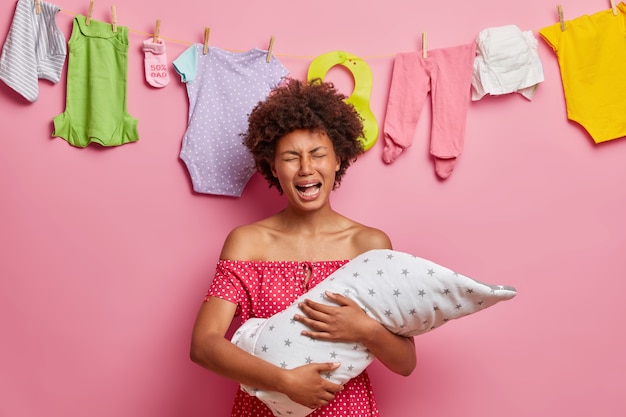 Depressed single mother being very tired of nursing baby, holds her newborn child in arms, cries from despair, sad to born sick infant, isolated over rosy wall, washed clothes on rope.