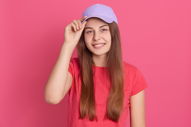 Free photo desirable young smiling student girl wearing red casual t shirt and baseball cap, being in good mood, keeping fingers on cap visor