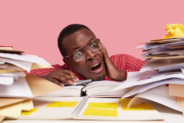 Desperate nervous dark skinned male student looks stressfully at pile of papers, writes information in notepad