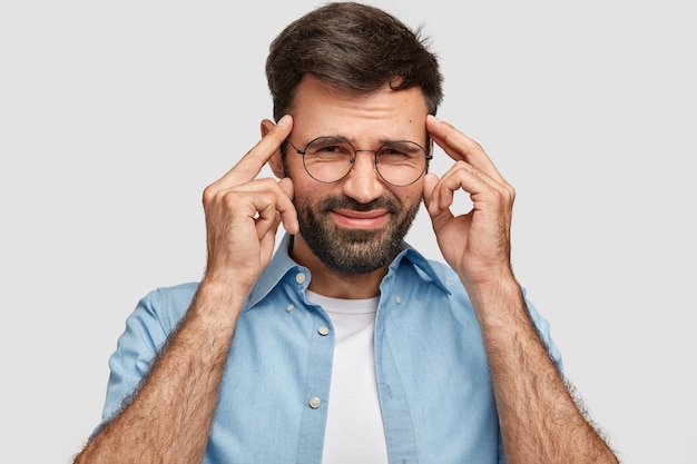 Desperate unshaven male keeps hands on temples, frowns face in displeasure, suffers from headache, dressed casually isolated over white wall. Handsome man expresses frustration, negative feeling