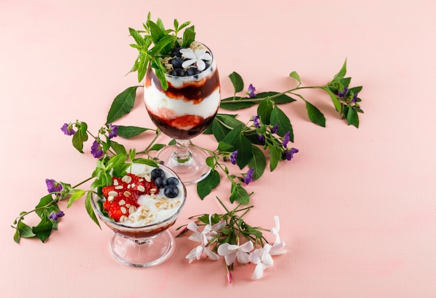 Free photo dessert with strawberries, blueberries, nuts, mint, flower branches in goblet and vase on pink surface, high angle view.