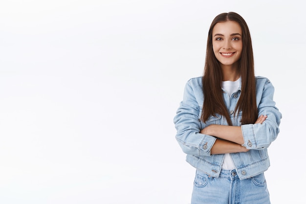 Determined, confident, happy brunette girl in denim jacket, jeans, cross arms over chest, smiling upbeat, looking assertive, self-assured, standing carefree over white background with friendly grin