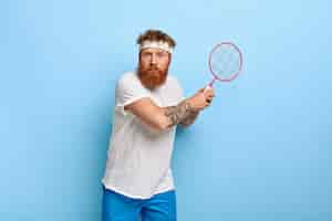 Free photo determined red haired tennis player holds racket while posing against the blue wall