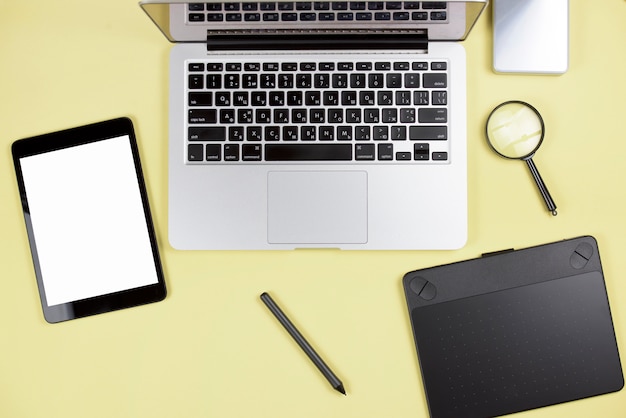 Digital tablet; stylus; graphic digital tablet; laptop and magnifying glass on yellow backdrop