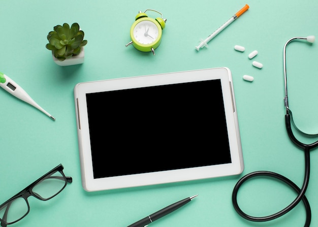 Digital tablet surrounded by medical equipment and alarm clock with succulent plant over green backdrop