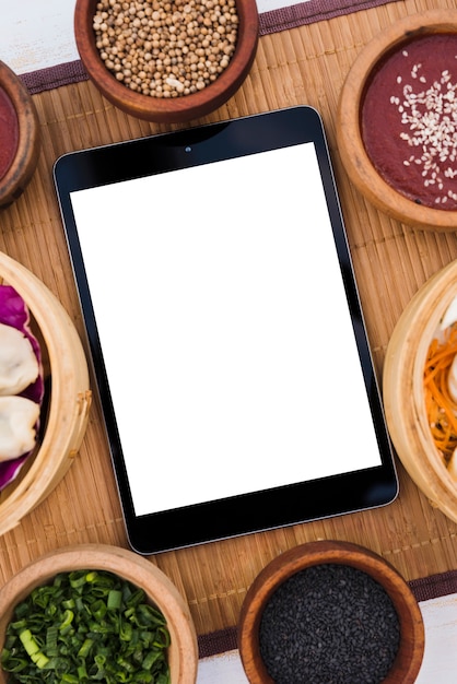 Digital tablet with white blank screen surrounded with steamers; coriander seeds; sesame seeds and spring onions on placemat