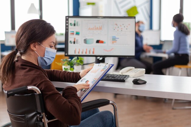 Disabled business woman with protective mask working in new normal business financial company typing on pc, checking reports