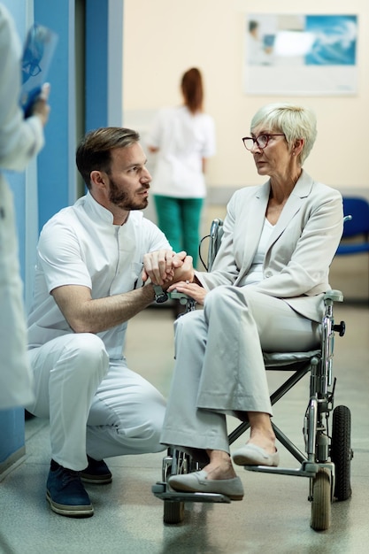 Free photo disabled mature woman in wheelchair talking with male doctor while holding hands in hospital hallway