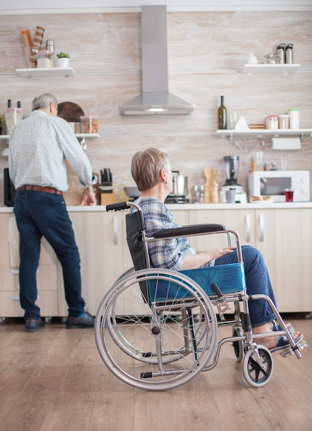 Disabled senior woman sitting in wheelchair in kitchen looking through window. Living with handicapped person. Husband helping wife with disability. Elderly couple with happy marriage.