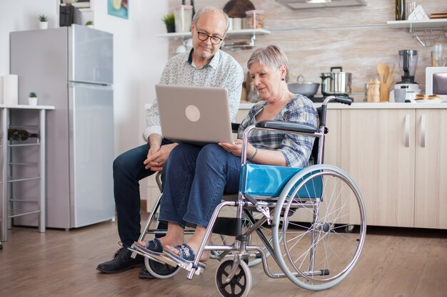 Disabled senior woman in wheelchair and her husband having a video conference on tablet pc in kitchen. Paralyzed old woman and her husband having a online conference.