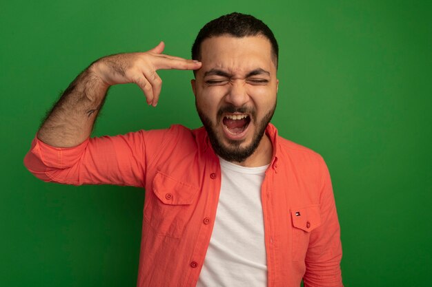 Disapointed young bearded man in orange shirt making pistol gesture near his temple with annoyed expression standing over green wall