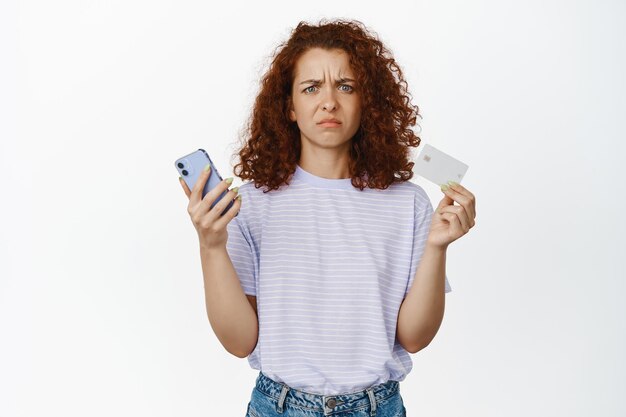 Disappoined, confused redhead girl holding credit card and mobile phone, frowning puzzled, dont understand, standing over white background.