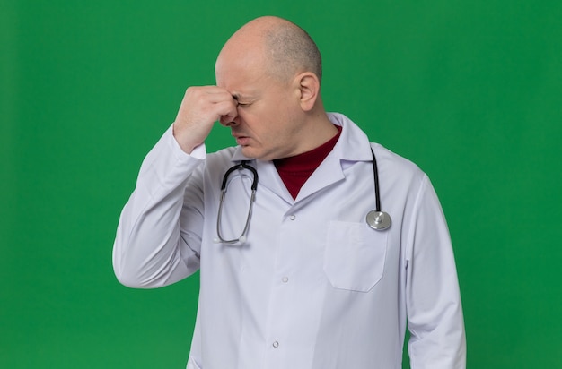 Disappointed adult man in doctor uniform with stethoscope holding his nose 