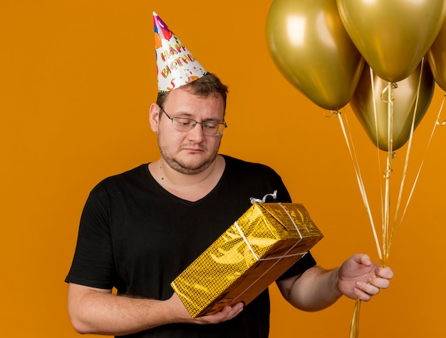 Disappointed adult slavic man in optical glasses wearing birthday cap holds helium balloons and gift box 