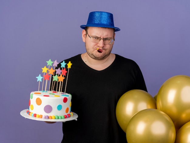 Disappointed adult slavic man in optical glasses wearing blue party hat stands with helium balloons holding birthday cake and blowing party whistle 