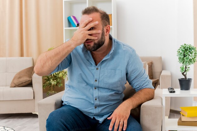 Disappointed adult slavic man sits on armchair putting hand on face looking at camera through fingers inside the living room