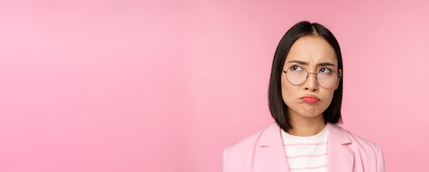 Disappointed asian businesswoman office lady in glasses looking upset at smth unfair sulking standing over pink background