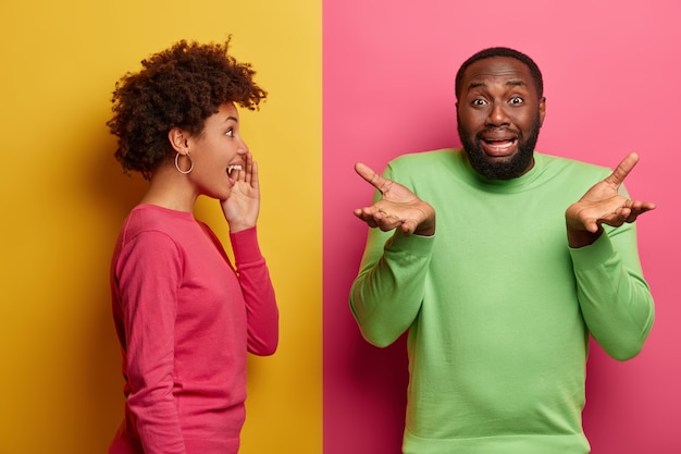 Free photo disappointed black man spreads palms, looks hesitant and unhappy, faces problematic situation, positive afro american woman whispers secret to boyfriend, stands sideways. pink and yellow color