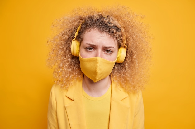 Free Photo disappointed displeased young woman wears protective mask feels lonely staying at home alone on self isolation listens music via wireless stereo headphones poses against yellow  wall