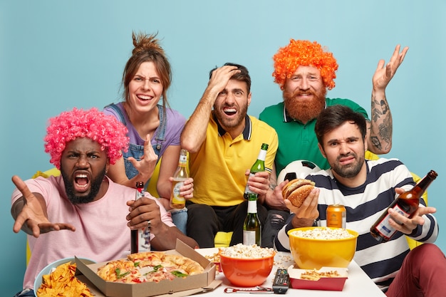 Disappointed four men and one woman watch sport game, dissatisfied with team failure, drink beer, have snack, express negative reaction, bad emotions, pose together on sofa at home. Team lose.