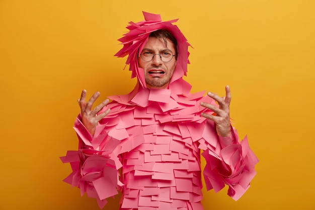 Disappointed frustrated man gestures and looks unhappily , finds out bad news, covered with pink sticky notes, cries from despair, isolated over yellow wall. Negative emotions