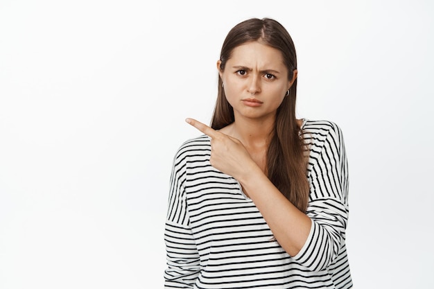 Disappointed girl pointing finger at upper left corner, showing smth bad, looking angry, standing over white background