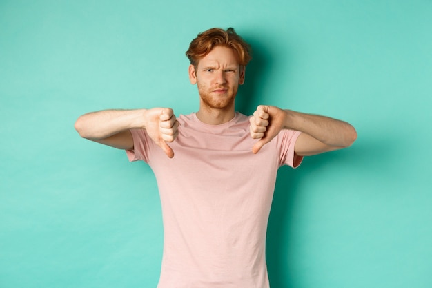 Free photo disappointed guy with red hair showing thumbs-down, frowning and looking skeptical, epxress dislike and disapproval, standing over turquoise background.