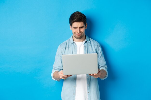 Disappointed handsome man looking at laptop screen and grimacing, judging something bad in internet, standing over blue background.