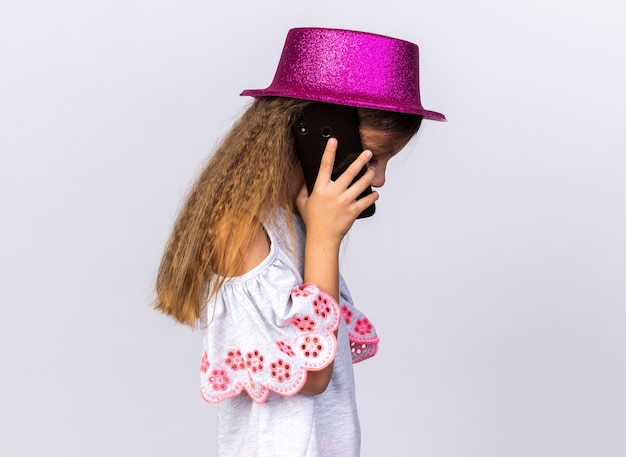 disappointed little caucasian girl with purple party hat standing sideways talking on phone isolated on white wall with copy space