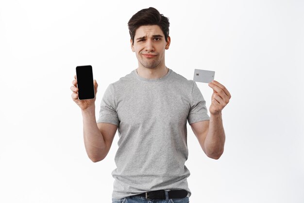 Disappointed man shows empty phone screen and credit card, displeased with online store, banking app service, frowning upset, standing over white background