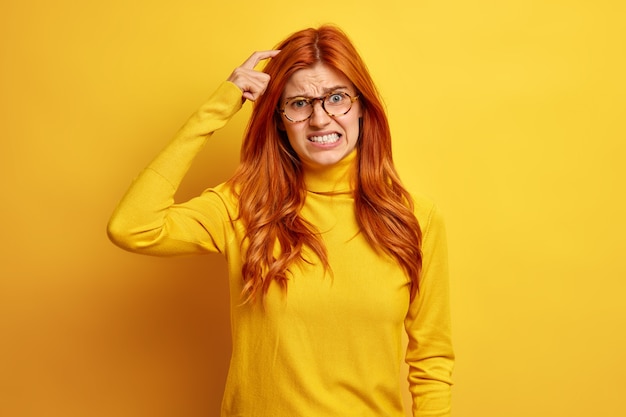 Disappointed redhead young woman has doubts scratches head and clenches teeth wears casual turtleneck has bad memory.