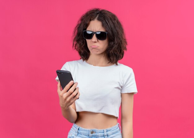 Disappointed young beautiful woman wearing sunglasses and holding mobile phone on isolated pink wall with copy space