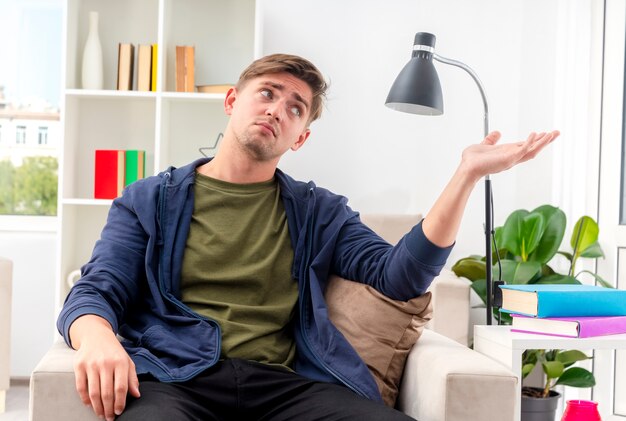 Disappointed young blonde handsome man sits on armchair looing and pointing at side with hand