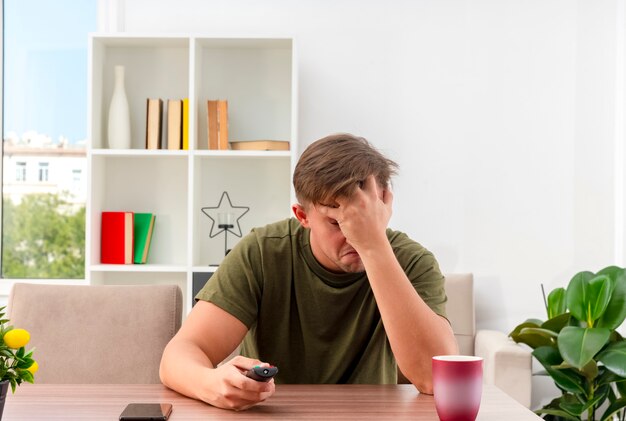 Disappointed young blonde handsome man sits at table with phone and cup