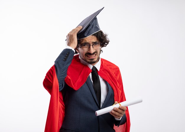 Disappointed young caucasian superhero man in optical glasses wearing suit with red cloak and putting hand on graduation cap holding diploma 