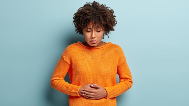 Free photo discontent afro american female suffers from abdominal stomach pain, touches belly with both hands, looks down, wears orange jumper, has menstruation, curly haircut, models over blue wall.