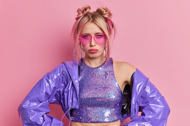 Free photo discontent blonde fashionable woman comes on eighties disco party dissatisfied with music looks sadly at camera wears trendy pink shades stylish purple clothes