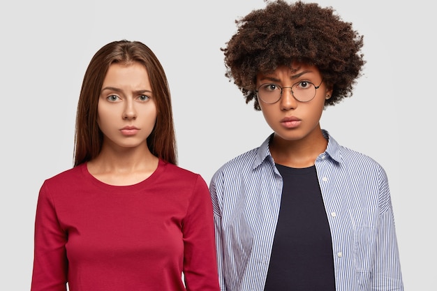 Discontent mixed race women frown faces in displeasure, stand closely to each other