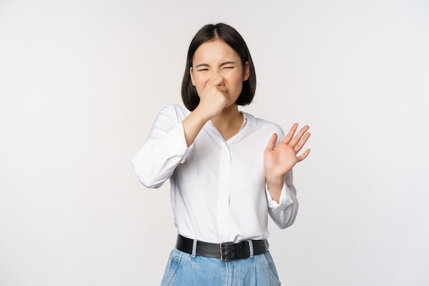 Disgusted asian girl close nose and showing rejection gesture digusting bad smell standing over white background