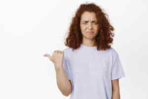 Free photo disgusting. displeased young redhead woman grimaces, points left with aversion and dislike, stands in tshirt against white background