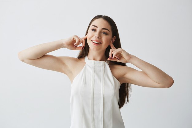 Displeased beautiful brunette girl closing ears with fingers from noise smiling with opened mouth .