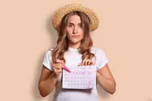 Free photo displeased caucasian female in casual t shirt and straw hat, indicates at period calendar, doesnt want to have menstruation during rest at seaside, isolated on beige. unhappy woman indoor