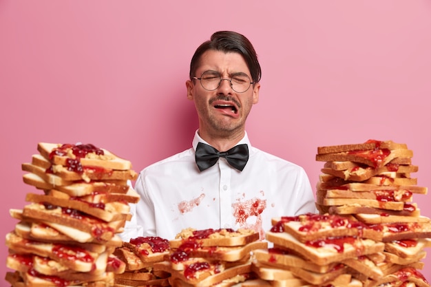 Displeased crying man has dirty white shirt as ate bread snack not carefully, expresses negative emotions, wears elegant clothes, has unlucky day, visits cafe or restaurant, isolated on pink wall