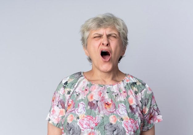 Displeased elderly woman yells with closed eyes isolated on white wall