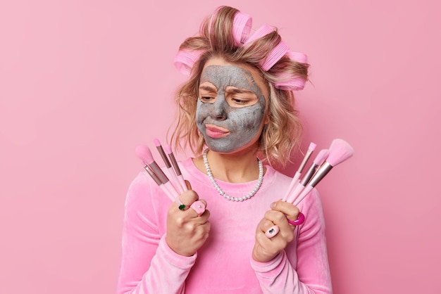 Free Photo displeased european woman applies clay mask on face chooses cosmetic brush going to do makeup wears hair rollers for making hairstyle dressed in festive outfit isolated over pink background