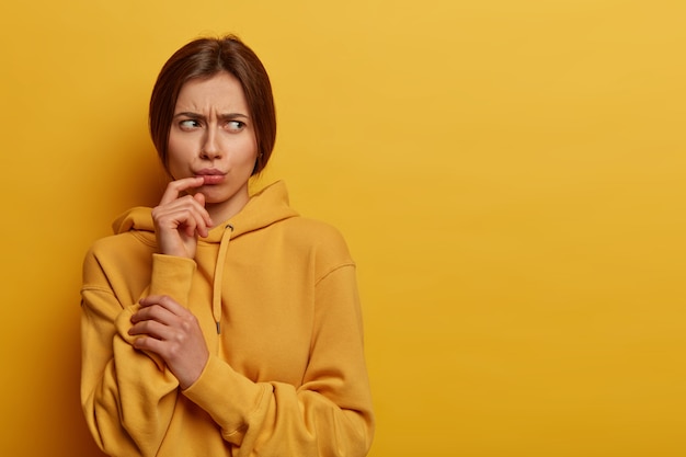 Free photo displeased gloomy young woman thinks about something troublesome and upsetting, looks angrily aside, pouts lips, frowns face, dressed in casual hoodie, isolated on yellow wall. negative feelings