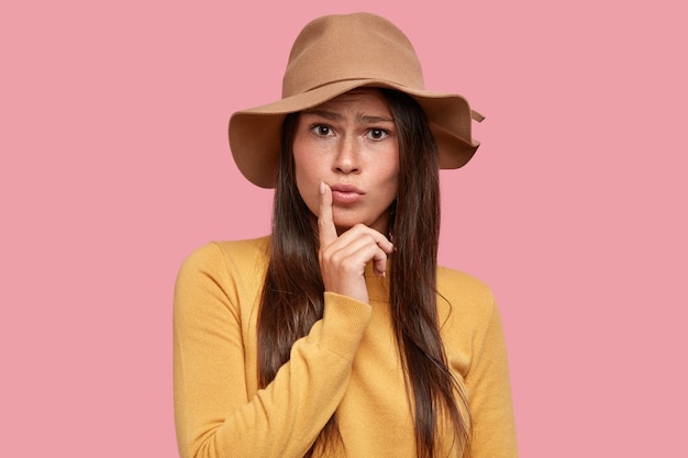 Displeased indignant teenager pouts lips, keeps finger near mouth,  casual sweater, look with puzzlement directly at camera