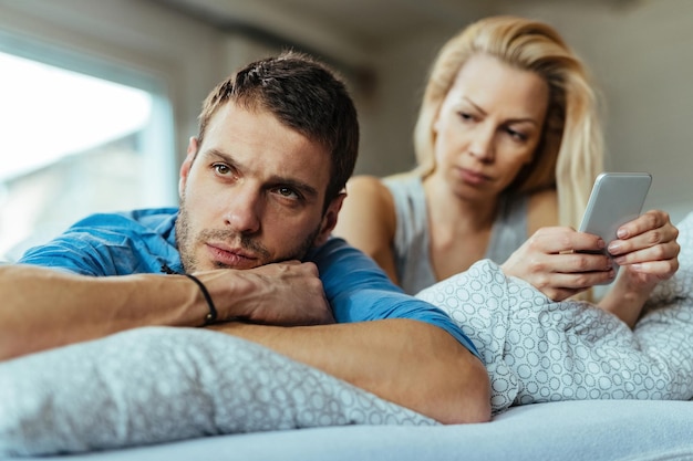 Free photo displeased man lying on the bed and ignoring his girlfriend who is text messaging on mobile phone