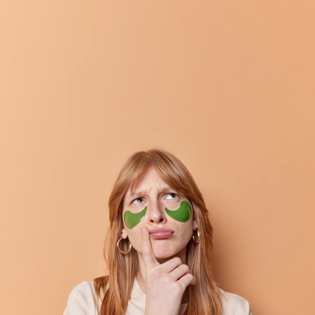 Free photo displeased red haired girl frowns and looks above keeps finger near lips thinks about something applies beauty patches for skin care isolated over brown background blank copy space overhead