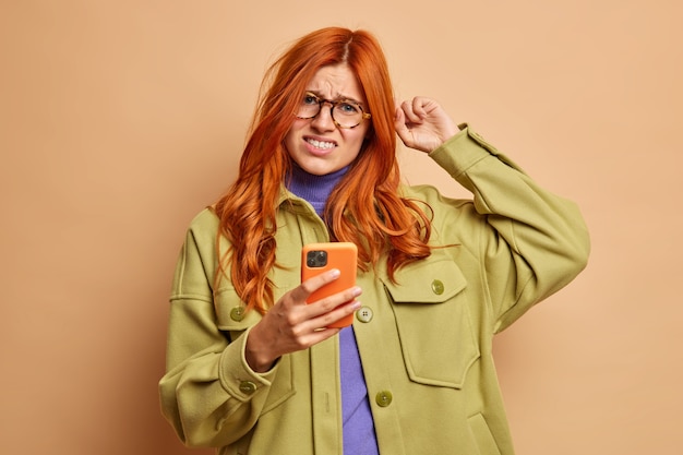 Free photo displeased red haired woman scratches head frowns from dissatisfaction tries to solve problem with smart phone doesnt know how to use new application dressed in fashion clothes.